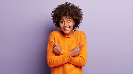 Indoor shot of cheerful woman points at different sides, feels enthusiasm and uncertainty, dressed in casual clothes, stands between choices, decides which deal to take, isolated over purple wall - obrazy, fototapety, plakaty