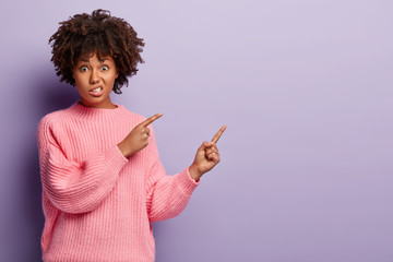 Displeased woman with annoyed facial expression, points aside with both fore fingers, dressed in oversized jumper, frowns face, has puzzled look, isolated over purple background. Look there.