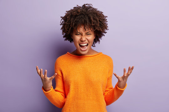 Angry annoyed female gestures with annoyance, screams loudly, expresses irritation, wears casual orange jumper, reproaches partner in betrayal, isolated over purple studio wall. Stop it please