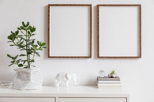 Stylish white home decor of interior with two brown wooden mock up photo frames with books, beautiful plant in stylish pot, elephant figure and home accessories. Minimalistic scandinavian room.