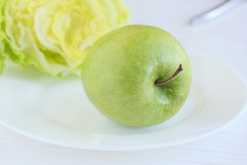 Juicy green organic apple with fresh green salad with selective focus at white plate on blurred neutral background. Green diet vegetarian ingredients. Healthy natural food for weight loss 