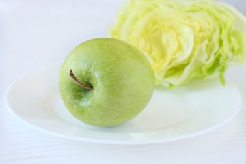 Juicy green organic apple with fresh green salad with selective focus at white plate on blurred neutral background. Green diet vegetarian ingredients. Healthy natural food for weight loss 