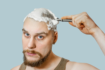 A guy with a beard and a razor in his hand, with foam and gel on his face shaves in the morning at...