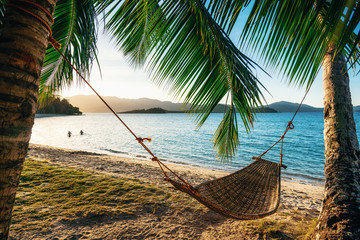Empty hammock between two palm trees on the beach at sunset. Silhouette of couple in the background in sea. Holiday and vacation concept - 252457218