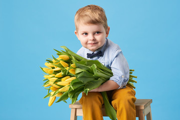 Adorable smiling child with spring flower bouquet looking at camera isolated on blue. Little toddler boy holding yellow tulips as gift for mom