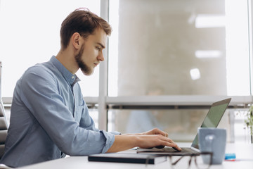 Full concentration at work. Handsome young beard man in shirt working on laptop while sitting at his working place