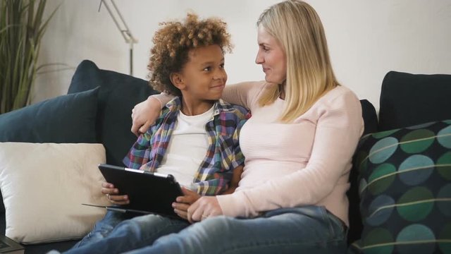 American woman and multi ethnic boy is sitting on sofa together and watching online using tablet.Multiracial male work with real digital tachnology app for relaxing exited time. Internet generation