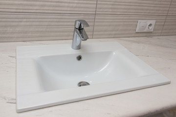 Ceramic white washbasin with stainless faucet mounted on a marble worktop against a background of gray large wall ceramic tiles. Bathroom interior element, close-up