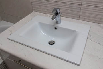 Ceramic white washbasin with stainless faucet mounted on a marble worktop against a background of gray large wall ceramic tiles. Bathroom interior element, close-up