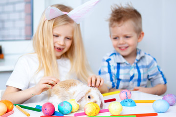 Blonde girl in rabbit ears on head and boy with little bunny. Colorful eggs and markers on table. Prepearing for Easter, family and holidays concept.