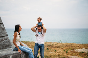 Happy young family in white t-shirts and blue jeans with a small daughter in dress sitting near to the lighthouse and playing