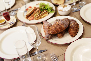 Breakfast table setting arrangement with bread, croissant, cappuccino and orange juice served as the menu. Illuminated with a warm morning light.
