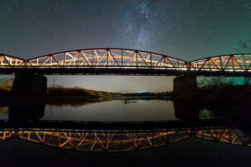 Illuminated metal bridge on concrete supports reflected in water on dark starry sky with Milky Way constellation background. Night photography concept.