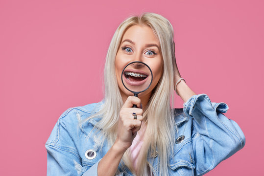 Happy joyful blond Woman in casual clothes wear brace and take magnifying on pink background. Broad smile