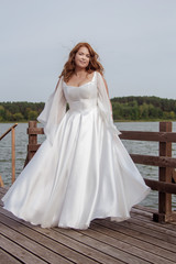 Red-haired bride in a white dress posing on a quay near a lake