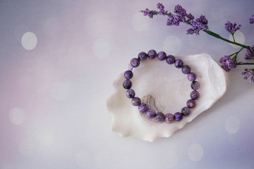 An close-up of an amethyst bracelet on a seashell next to a purple flower with a purple and white bokeh background. - obrazy, fototapety, plakaty