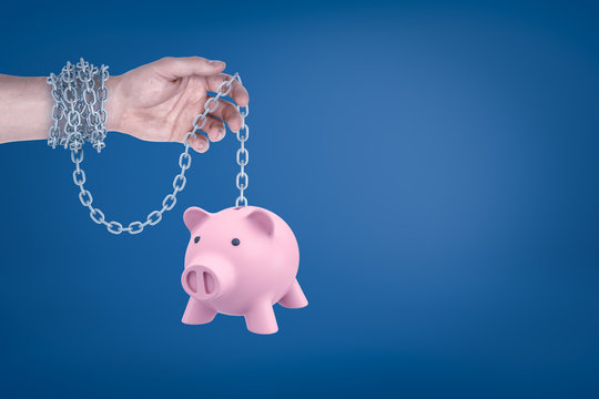 Close-up of man's hand with metal chain around wrist holding piggy bank hanging on the other end of chain.