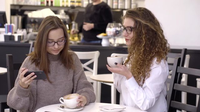 two friends with glasses are sitting in a cafe and taking selfies, smiling, looking at photos on the phone.