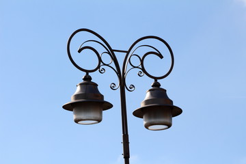 Baroque style dual street lamps held together by dark metal support on clear blue sky background