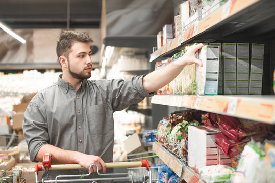 Portrait Of A Man With A Beard Stands With A Cart On The Aisle In The Supermarket And Takes Cereal Breakfast From The Shelf. Buyer Chooses A Box Of Flakes On The Grocery Store Shelf.