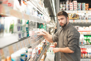 Portrait of amazed man standing near the refrigerator in the milk department of the supermarket and looking at the camera. Buyer chooses yogurt in a grocery store, looks at the camera with amazement.