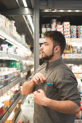 Happy man wearing a shirt stands at the refrigerator in the milk department of the supermarket and takes yogurt from the shelf. Buyer with a beard takes a yogurt from his fridge at a grocery store.