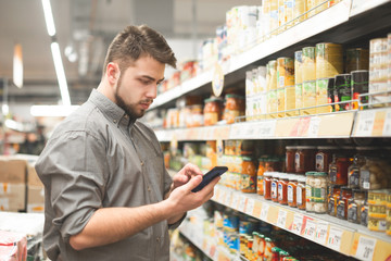 Man stands in the aisle of the supermarket at shelves with canned vegetables and uses a smartphone. Buyer man looks at the shopping list on the smartphone and selects the products in the supermarket