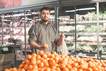 Happy man buys tangerines in a supermarket, stands with a packet and citrus in his hands. Smiling...