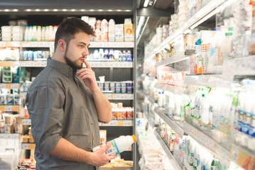Portrait of a pensive man standing in the supermarket dairy department, looks at shelves with bottles of milk and thinks. Buyer chooses which dairy product to buy.Adult man buys milk.