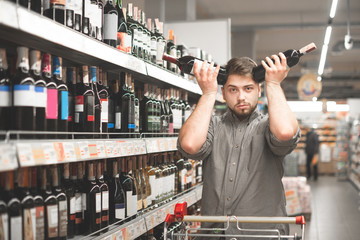 Portrait of a husband buys wine in a supermarket,holds two bottles and does not know what to choose, looks at the camera. Portrait of a buyer in a supermarket holds wine bottles over his head
