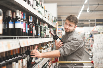 Сheerful man buys wine at a grocery store. Funny man with bottles of wine in the alcohol department of the supermarket. Positive buyer takes the wine out of the shelves and looks at the camera.