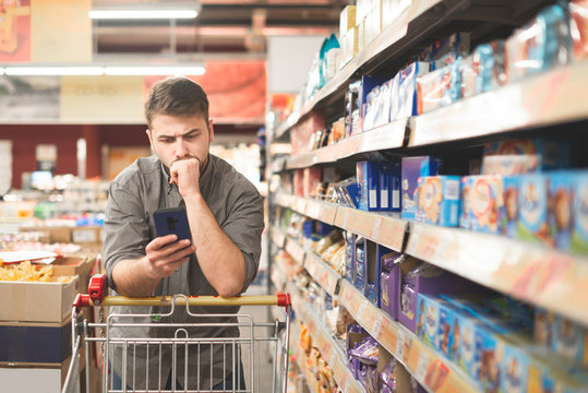 Serious man stands in a cart in the aisle of a supermarket, looking at the screen of a smartphone.Buyer in a supermarket uses a smartphone. Man in a smartphone in the hands of a grocery store