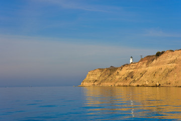 Fototapeta na wymiar White small lighthouse on cliff by blue Greek sea