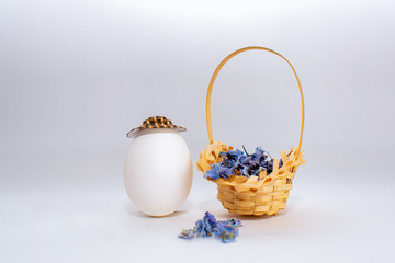 Easter egg in a hat and Wicker basket with blue wildflowers on white pastel background. Creative idea. Art food concept.