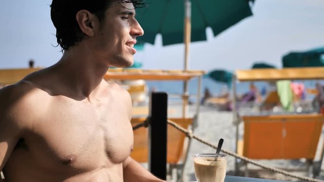 Athletic man at the seaside sitting at a beach bar with the sea behind him, smiling to camera