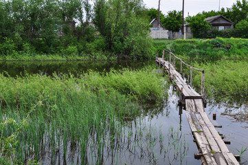 Fototapeta na wymiar Old small wooden bridge across the overgrown river in the village.