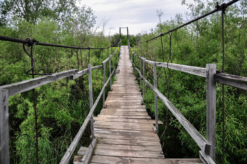 Old suspension wooden bridge across the overgrown river in the village.