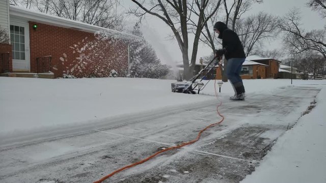 Man in black jacket with hood wearing tall boots and blue jeans clears from snow his walkway using corded electric push snowblower machine.
