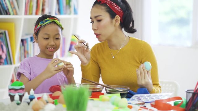 Cheerful little girl and her mother painting easter eggs together using dyes and paintbrush on the table at home. Shot in 4k resolution