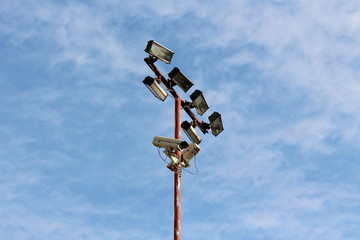 High metal pole with multiple light reflectors and security camera connected with electrical wires on cloudy blue sky background