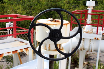 Large black metal valve on top of white pipeline surrounded with pipelines and platforms at...