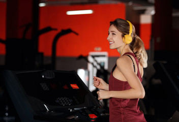 Fototapeta na wymiar Young woman with headphones listening to music and running on treadmill at gym