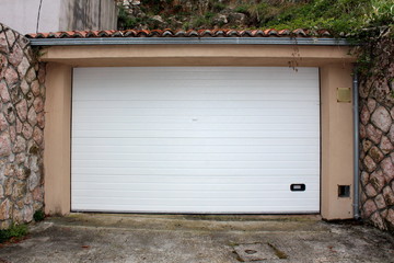 Modern white rollup garage doors with small handle for manual opening mounted on renovated garage wall surrounded with traditional stone wall and concrete driveway in front on warm sunny day