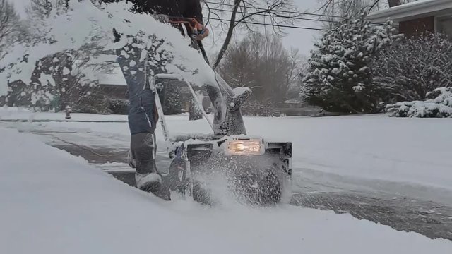 Super slow motion of electric snowblower equipped with light being pushed through sidewalk covered in thick layer of snow showing machine auger and auger in action.