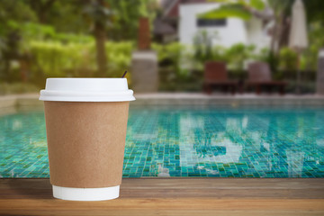 Fototapeta na wymiar PAPER COFFEE CUP on wood counter bar open space to outdoor blur swimming pool background. Coffee cup in front of blur green water in swimming pool. Close up and selective focus.
