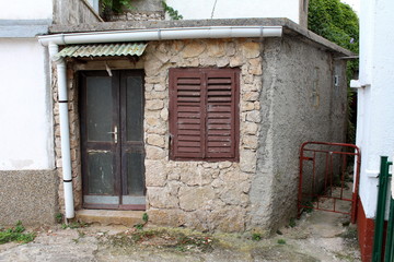 Fototapeta na wymiar Stone and concrete small house with dilapidated cracked wooden doors and closed window blinds with new white metal gutter surrounded with concrete path on warm cloudy day