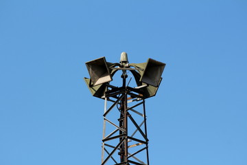 Top of tall metal structure holding four large public civil defence warning air sirens on clear blue sky background