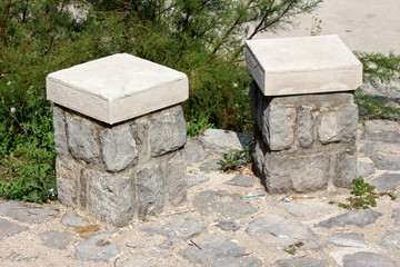 Two single seats stone and concrete public benches on stone tiles foundation with dense plants in...