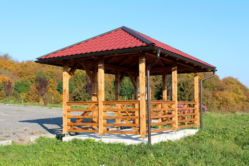 Wooden gazebo structure with new roof and gutter mounted on concrete foundation next to uncut grass and gravel parking with dense trees and clear blue sky background