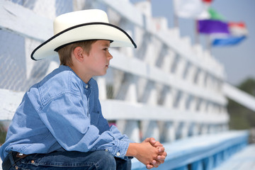 A young boy sitting on the bleachers at the county fair rodeo wearing a cowboy hat.